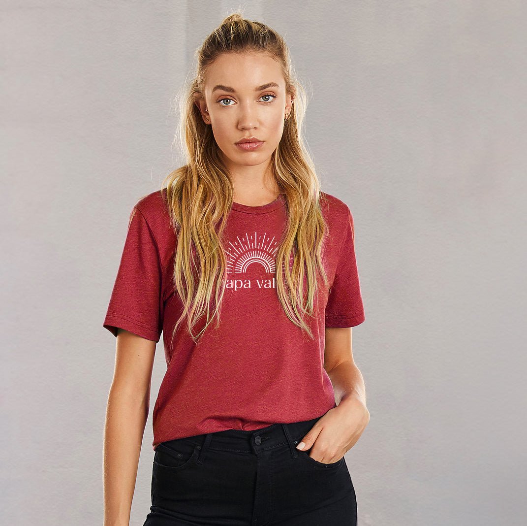Best-Selling Tees for Every Destination featuring a model wearing a burgundy Bella + Canvas unisex tee with a sun and rainbow "sunshine" design in front of a neutral background.