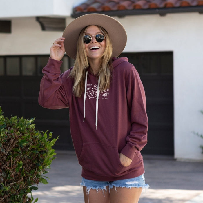 Woman wearing a maroon hoodie with Slant collection design and denim shorts standing in front of a white building with a tiled roof.